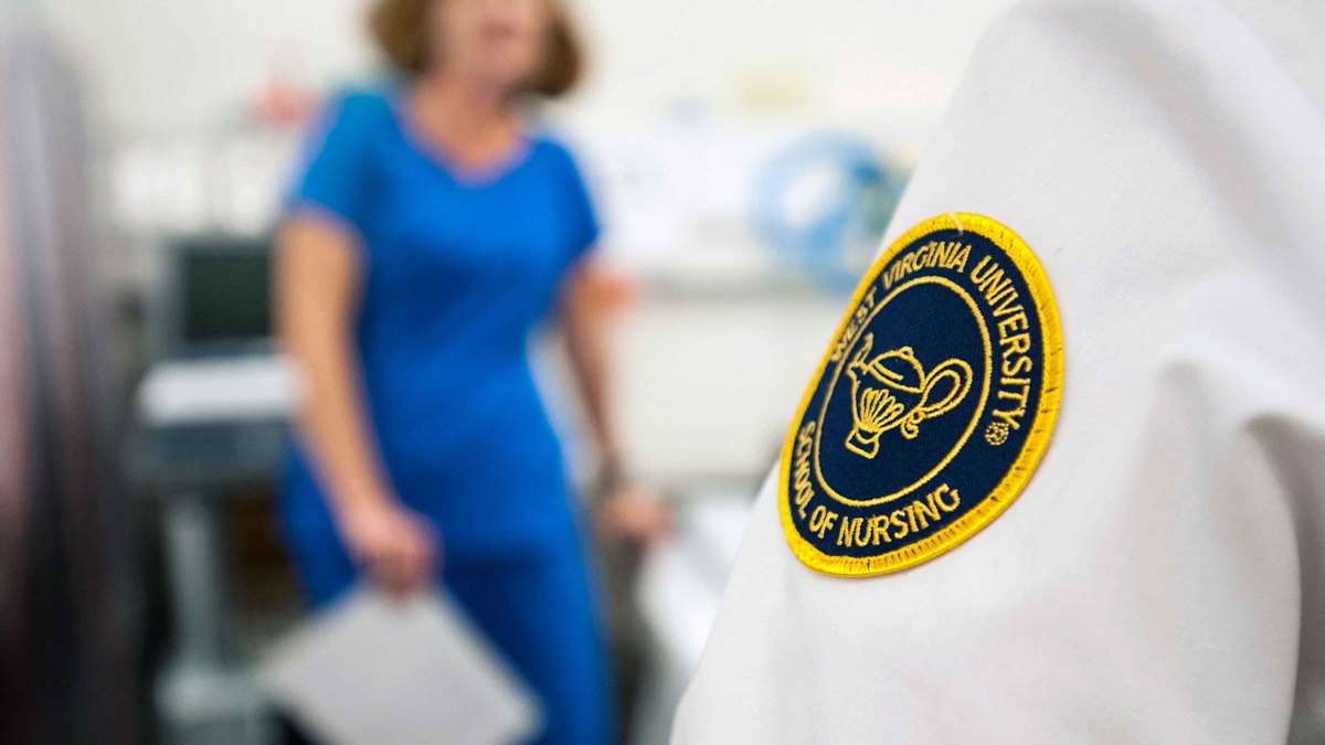 A photo of a nurse’s sleeve with the School of Nursing patch in the foreground, in front of an out-of-focus nurse standing in the background.