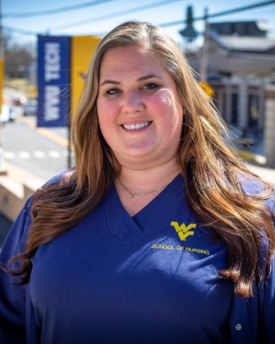Petra is smiling while wearing blue WVU School of Nursing scrubs. In the background behind her is a street scene with WVU Tech banners hanging on light poles.