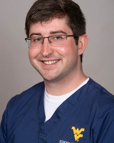 Ben is shown smiling, while wearing blue WVU School of Nursing scrubs.